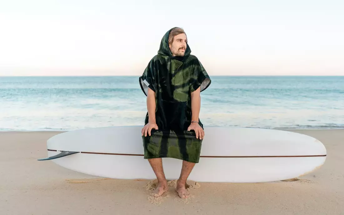 mann im gruen schwarz gemustertem surfponcho sitzen auf surfbrett vorm meer