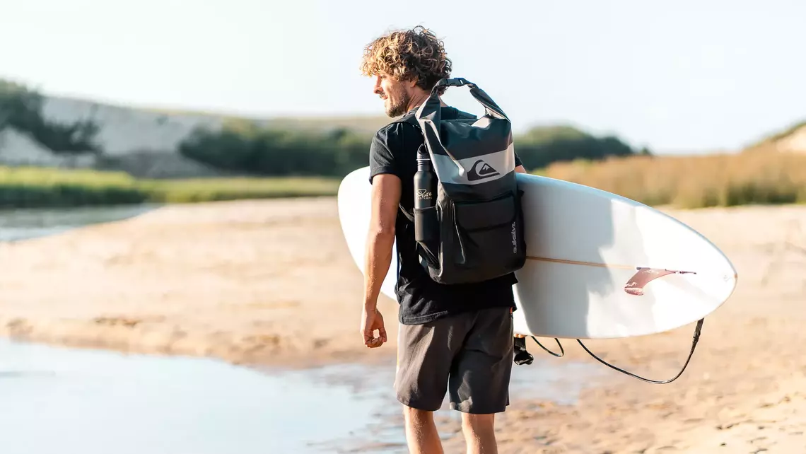 Surfer mit Surfrucksack und Surfbrett unter dem Arm am Strand