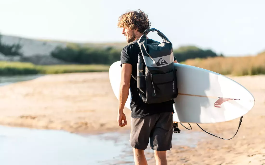 Surfer mit Surfrucksack und Surfbrett unter dem Arm am Strand